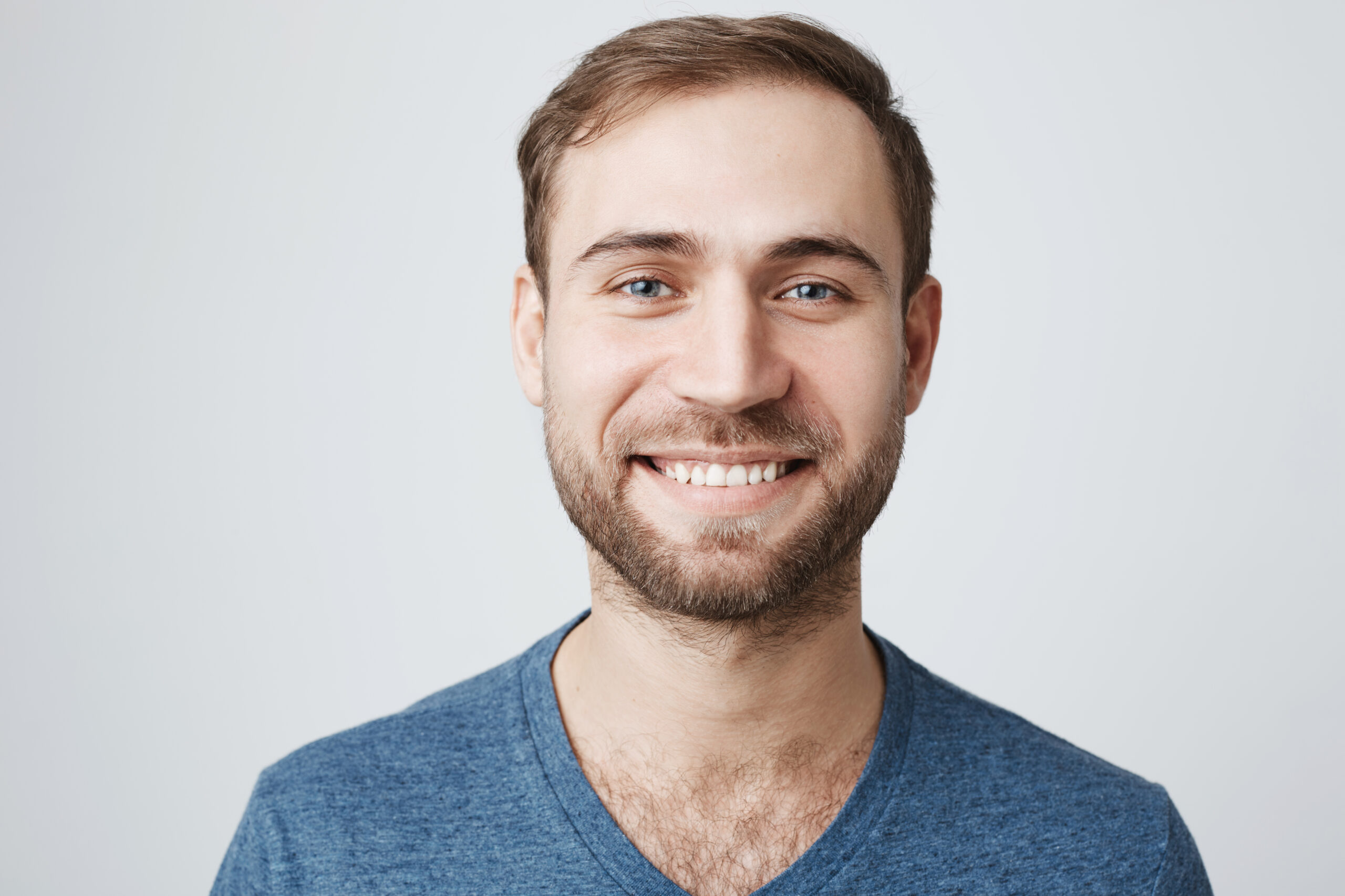 Pleased good-looking young man with beard and trendy hairdo wears casual blue t-shirt, smiles pleasantly at camera, glad to receive good news, isolated against gray background